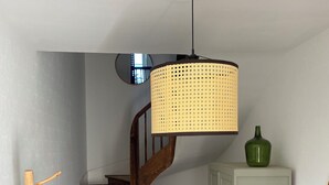 Una escalera con peldaños de madera y barandal negro, una lámpara colgante de ratán, un florero verde sobre un mueble y una silla de mimbre.