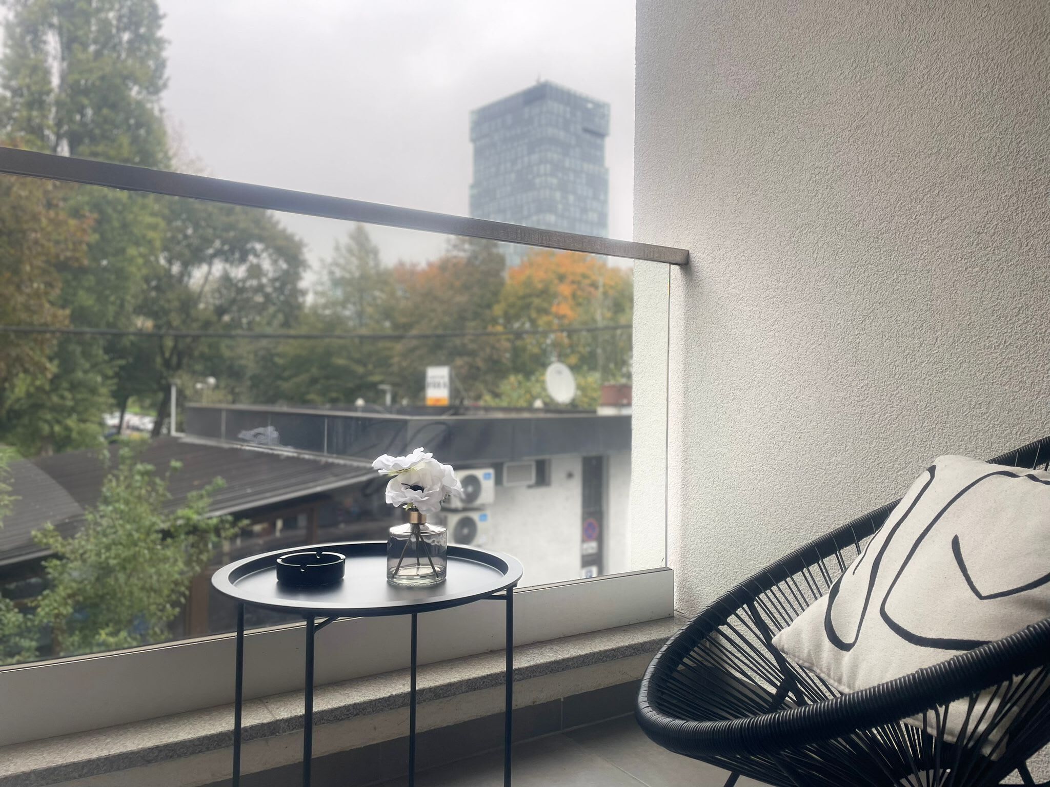 Ein Balkon mit Blick auf Bäume und ein Gebäude, ein kleiner Tisch mit Kerze und Schale sowie ein schwarzer Korbstuhl mit Kissen.