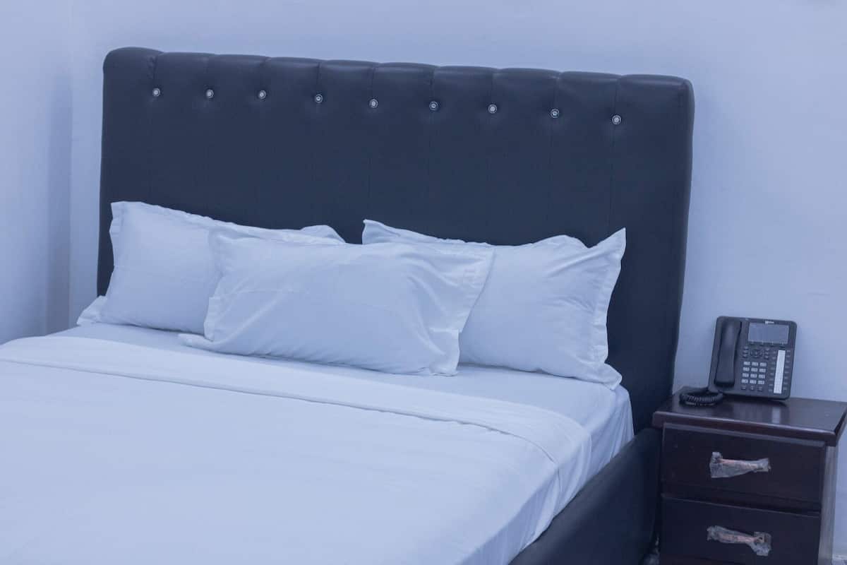 A bed with white bedding and a dark headboard. There is a bedside table with a telephone and two drawers.