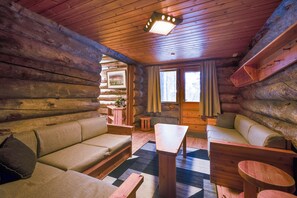 Interior de una cabaña de troncos con un sofá, una mesa y una ventana con cortinas.