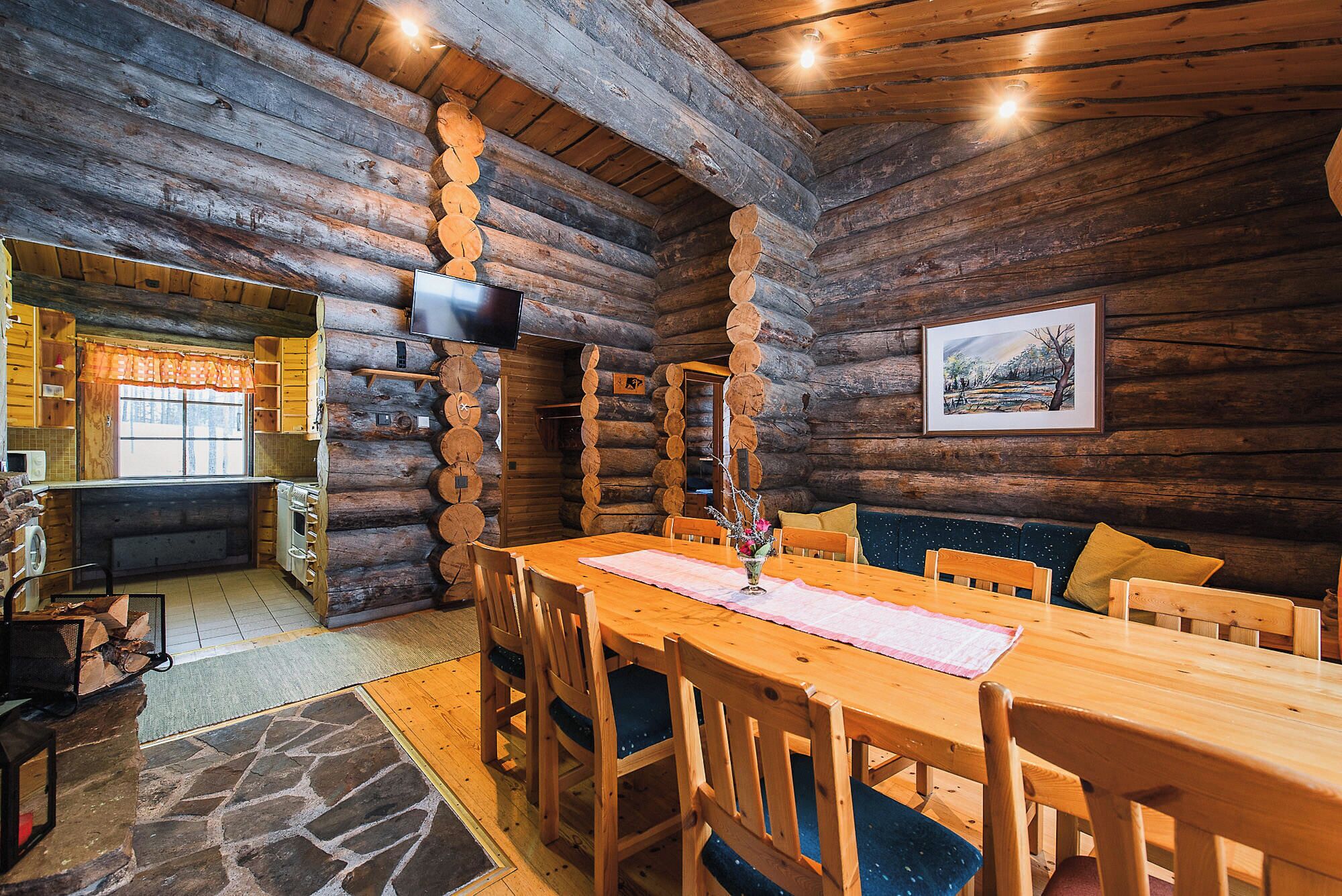 A log cabin interior with a wooden dining table, chairs, a fireplace, and a television mounted on the wall.