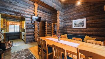 A log cabin interior with a wooden dining table, chairs, a fireplace, and a television mounted on the wall.