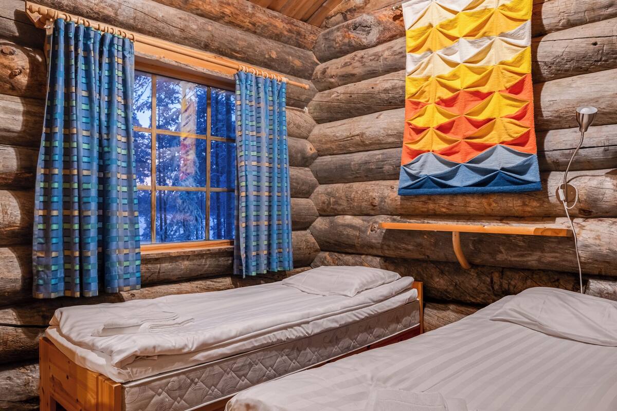 Quarto de cabana de toras com duas camas, uma janela com cortinas e um colcha colorida pendurada na parede.