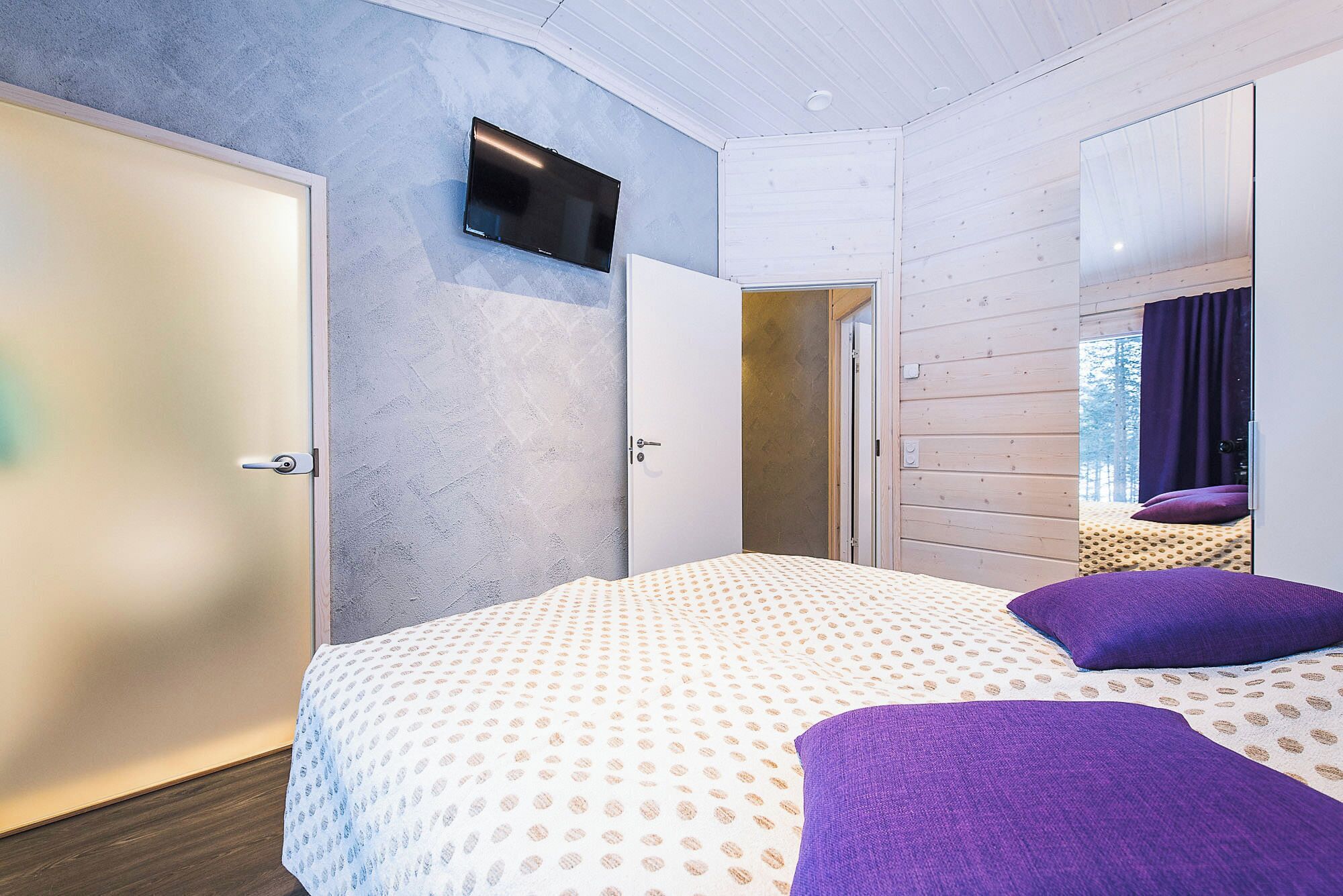 A bedroom with a bed, a TV mounted on the wall, and a door leading to another room.