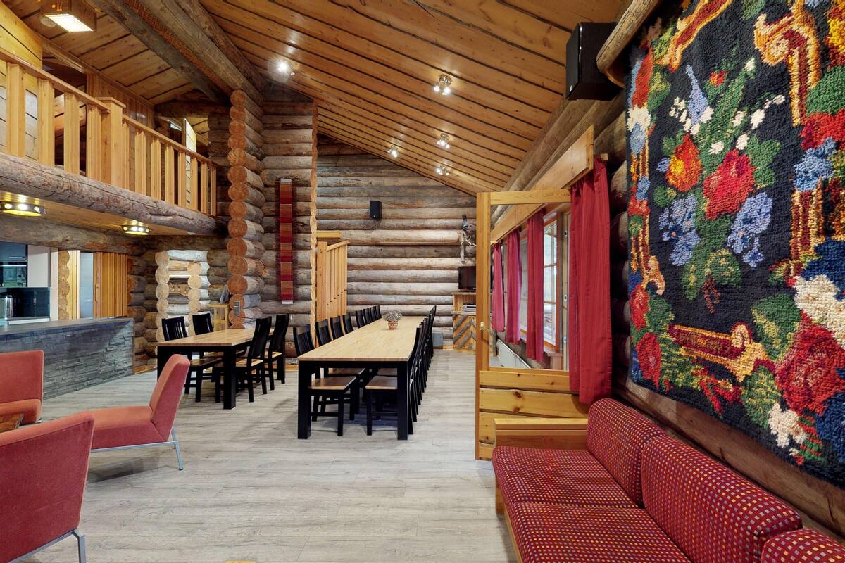 Interior de uma cabana de madeira com uma longa mesa de jantar, cadeiras estofadas em vermelho e uma grande tapeçaria na parede.