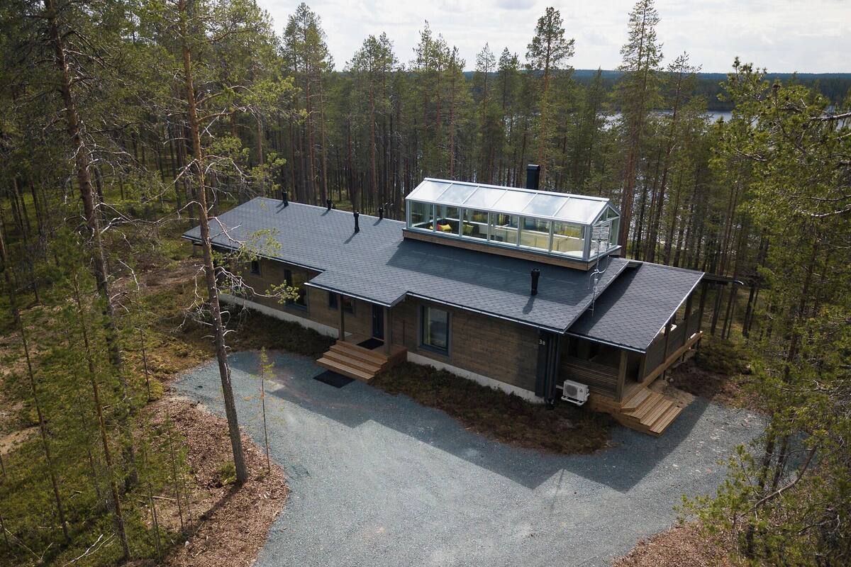 Uma casa moderna de dois andares com uma grande estufa de vidro no andar superior, cercada por uma área arborizada.