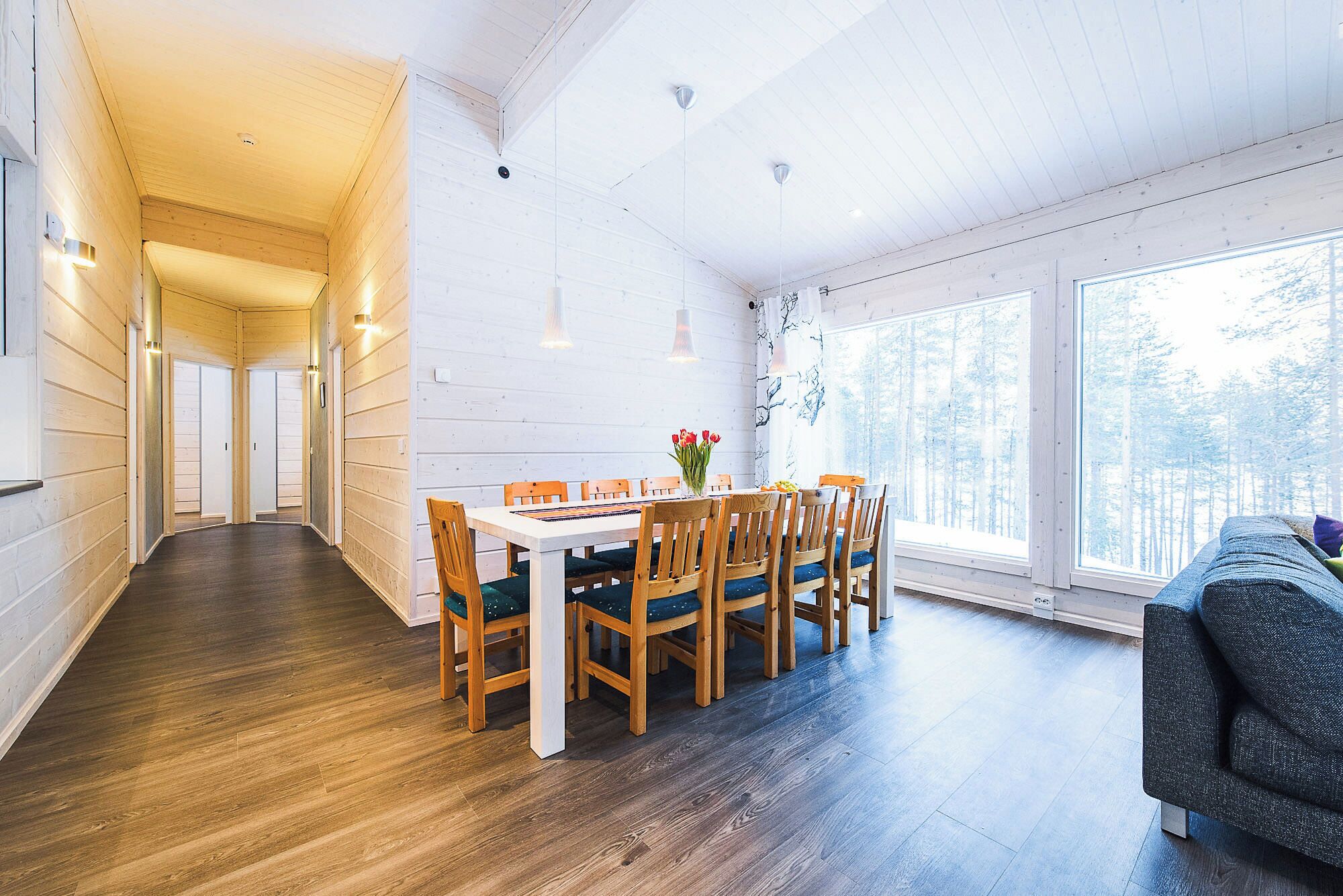 A spacious dining area with a long table and chairs, large windows with a view of a forest, and wooden walls.