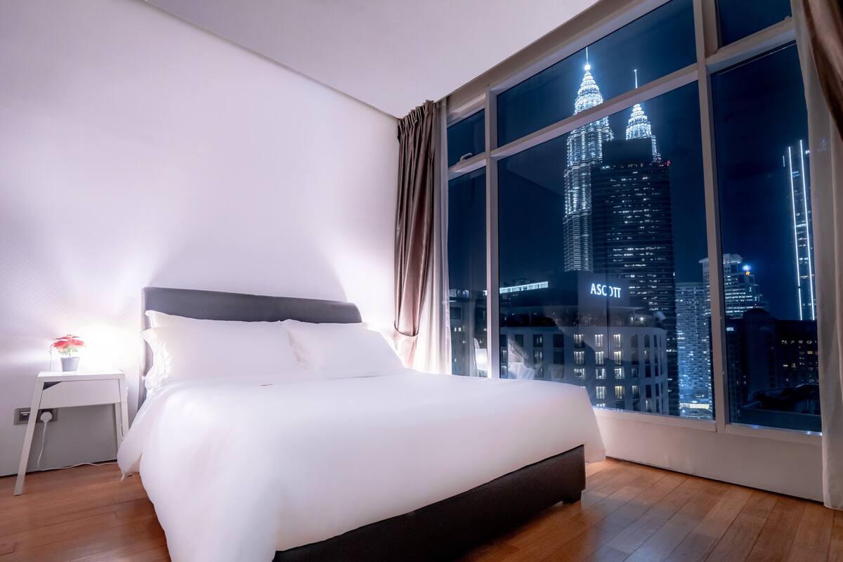 A hotel room with a large window offering a nighttime view of a city skyline, including a prominent illuminated tower.
