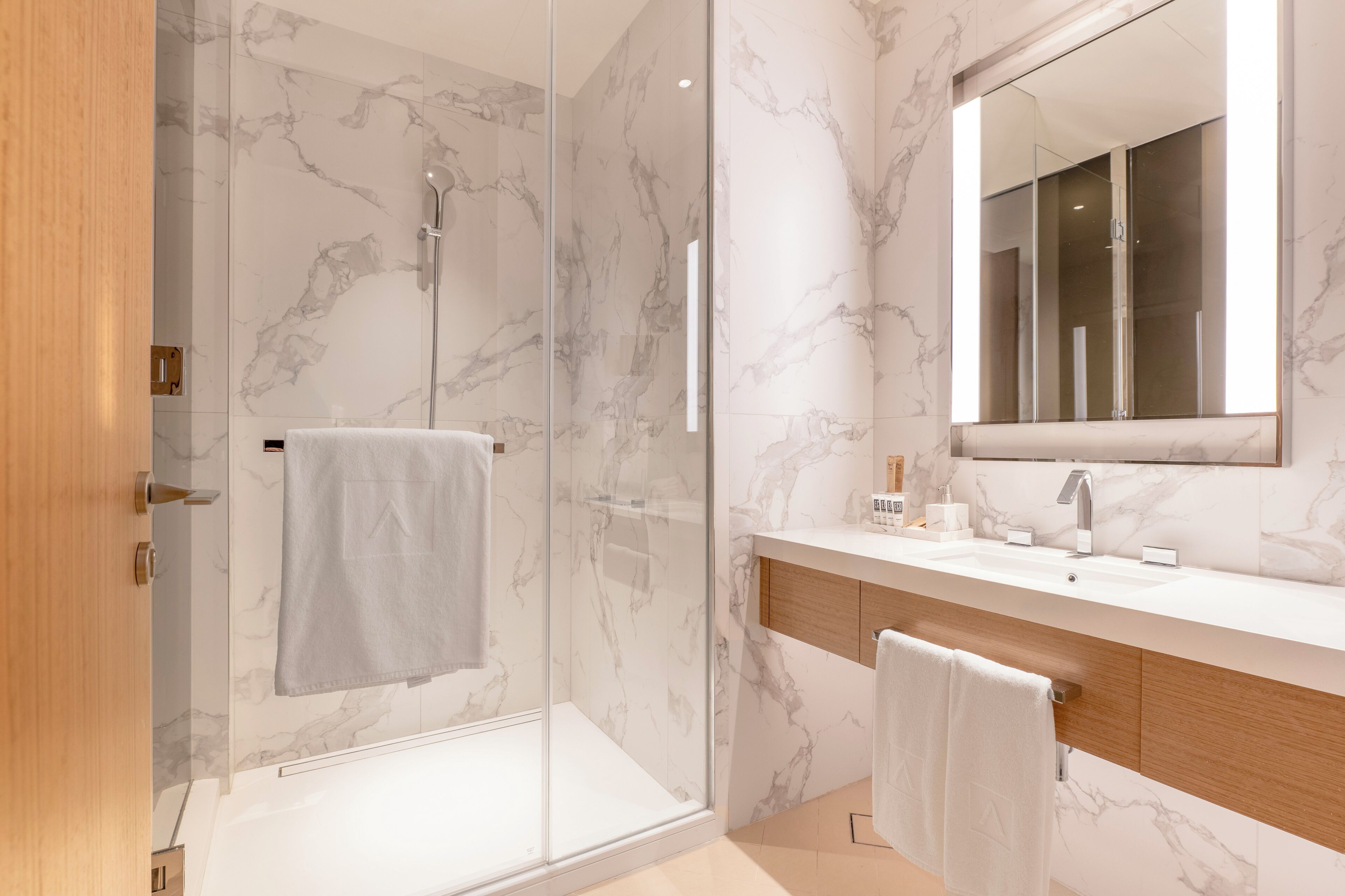 Une salle de bain moderne dotée d’une douche à parois de verre, d’un mur recouvert de carreaux de marbre, d’un grand miroir et d’un meuble-lavabo en bois.