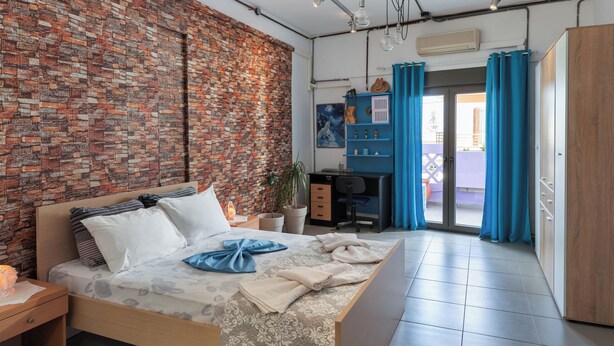 A modern bedroom with a brick wall, a bed with a patterned bedspread, a desk, and a wardrobe. - THENA apartment in  Limenas Chersonisou (Limenas Chersonisou)