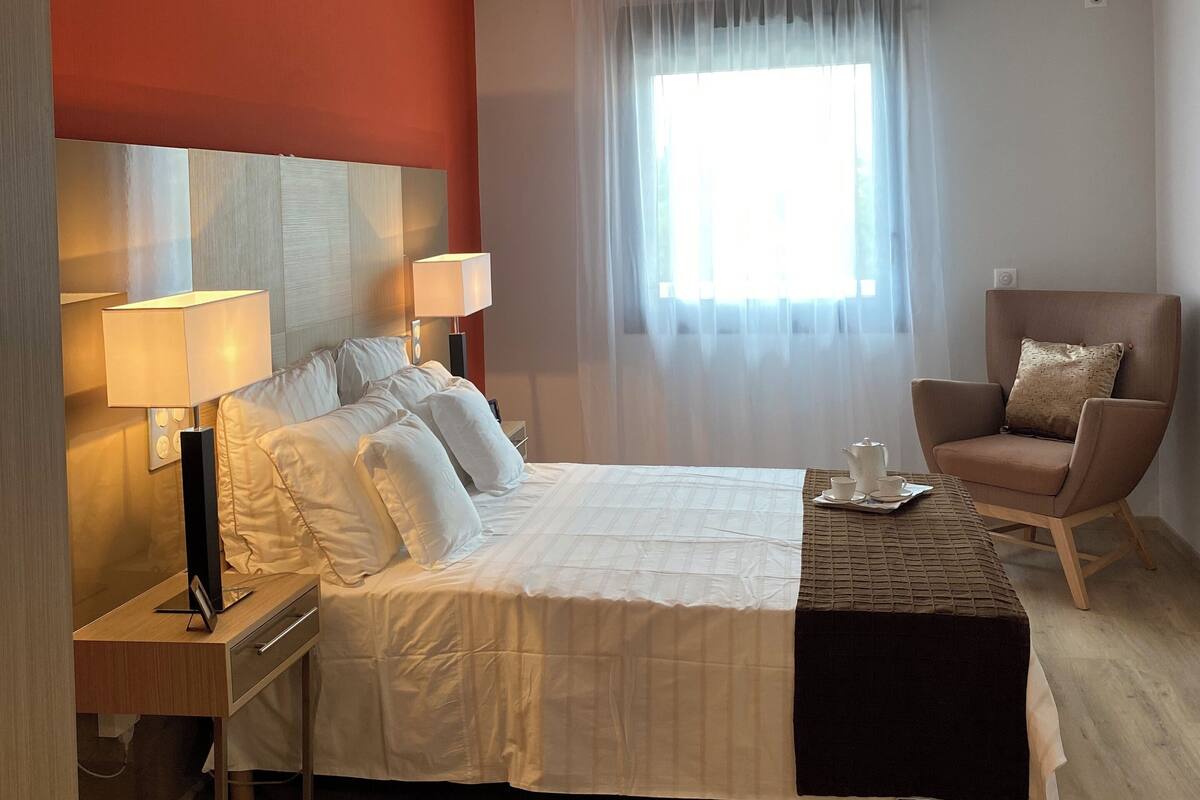 Une chambre d’hôtel avec un lit, des tables de chevet, une chaise, une fenêtre avec des rideaux légers et un luminaire au plafond.