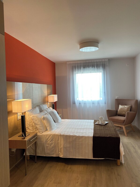 Une chambre d’hôtel avec un lit, des tables de chevet, une chaise, une fenêtre avec des rideaux légers et un luminaire au plafond.