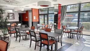 A modern restaurant with a clean, organized layout featuring multiple dining tables and chairs, large windows offering a view of the outside, and a mix of natural and artificial lighting. - Appart One Le Cannet (Le Cannet)