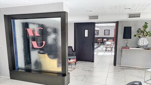 A modern hotel lobby with a large glass wall displaying a logo, a reception desk, and a seating area with a plant. - Appart One Le Cannet (Le Cannet)