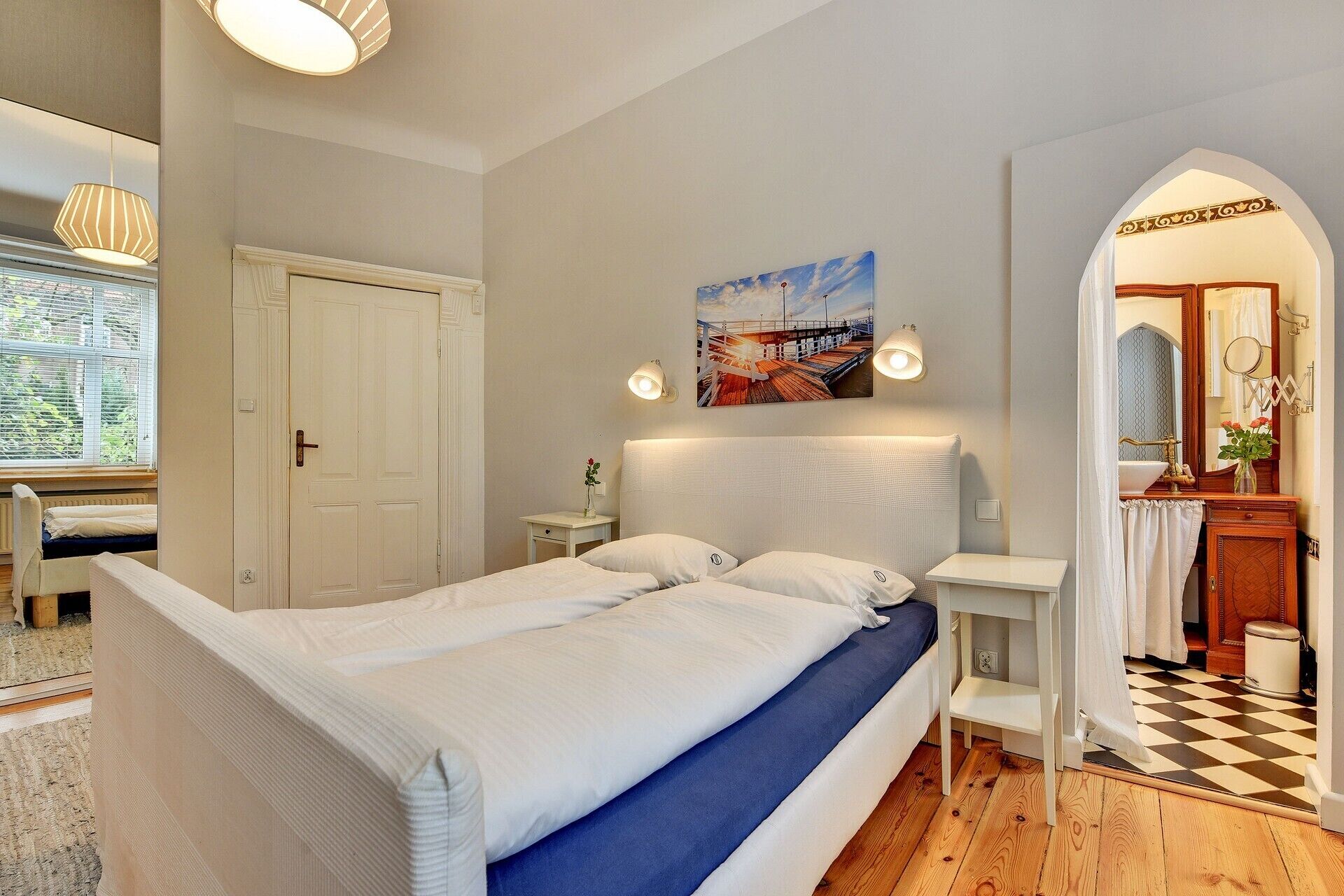 Een slaapkamer met een groot bed, een klein nachtkastje, een raam met jaloezieën, een deur en een schilderij aan de muur.