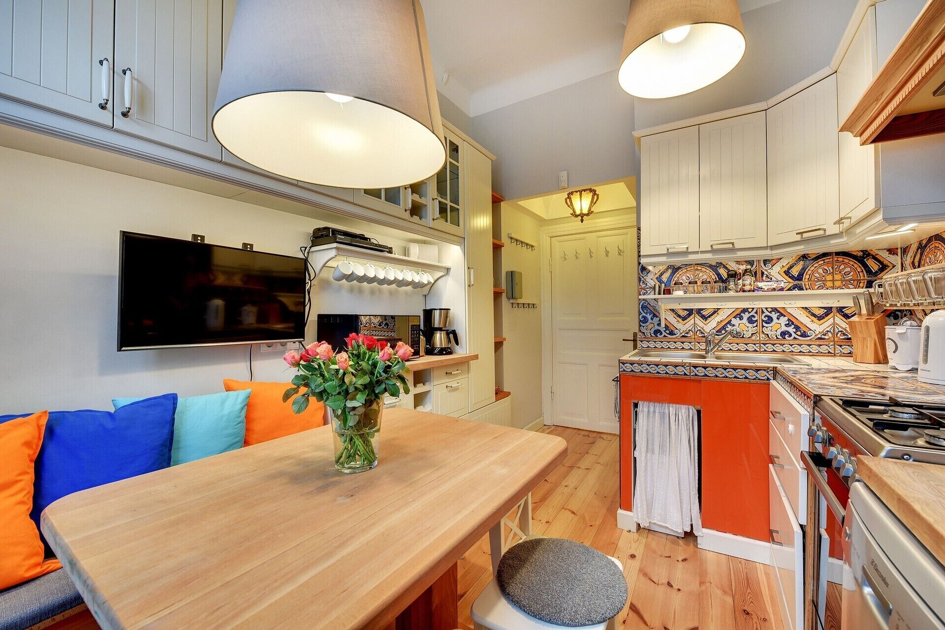 A modern kitchen with white cabinets, a wooden dining table, and a TV mounted on the wall.