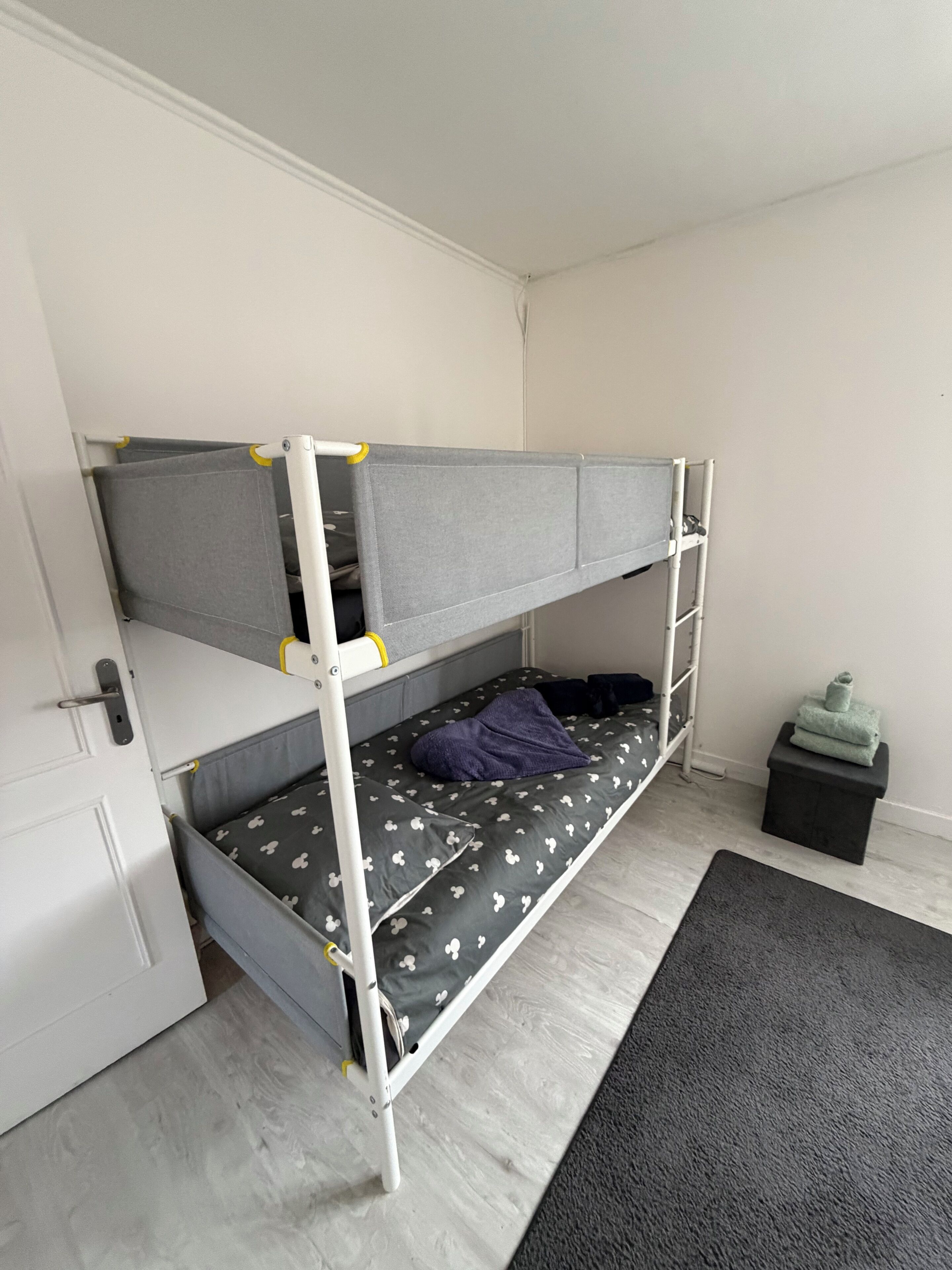 A bunk bed with a grey mattress and patterned pillow, a small black stool, and a grey rug on a light wooden floor.