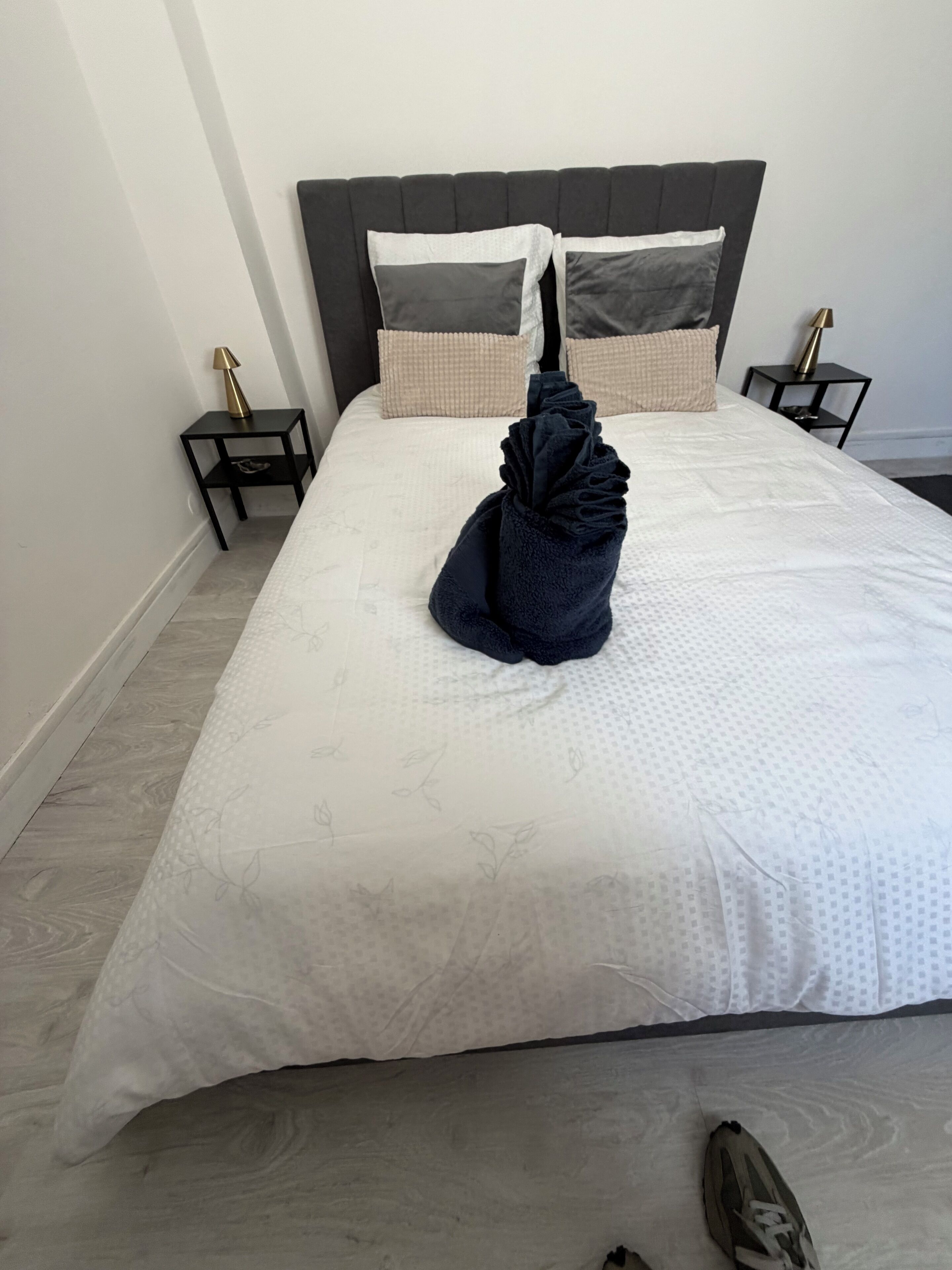 Ein ordentlich bezogenes Bett mit weißer Bettdecke und einem dunkelblauen, gefalteten Handtuch. Zwei Nachttische mit Lampen stehen bereit, und der Raum hat ein schlichtes, aufgeräumtes Design.