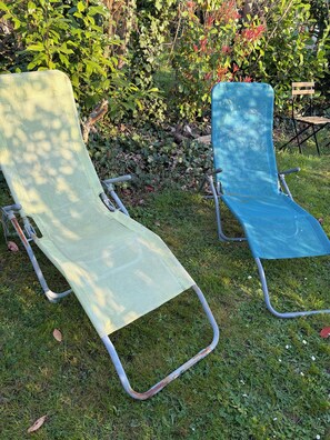 Two outdoor lounge chairs with metal frames and fabric seats, positioned on grass with a garden and wooden bench in the background.