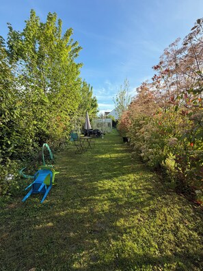 Un jardín con césped, un invernadero, una mesa con sillas y una silla azul y verde.