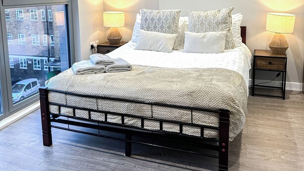 A modern bedroom with a large bed, two bedside tables with lamps, a round mirror on the wall, and a view of the city through a large window. - 3-Bed Hoxton Flat w/ Private Balcony & Lift access near Shoreditch , Old Street (London)