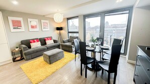 A modern living room with a grey sofa, ottoman, and black chairs. There is a yellow rug, a floor lamp, and a dining table with chairs. The room has large windows with a city view. - 3-Bed Hoxton Flat w/ Private Balcony & Lift access near Shoreditch , Old Street (London)