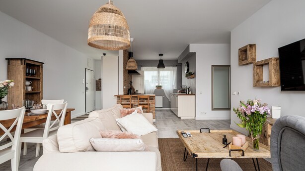 Ein modernes Wohnzimmer mit einer Couch, einem Couchtisch und einem Essbereich. Im Hintergrund befindet sich eine Küche mit einem Fenster und einer Hängelampe.