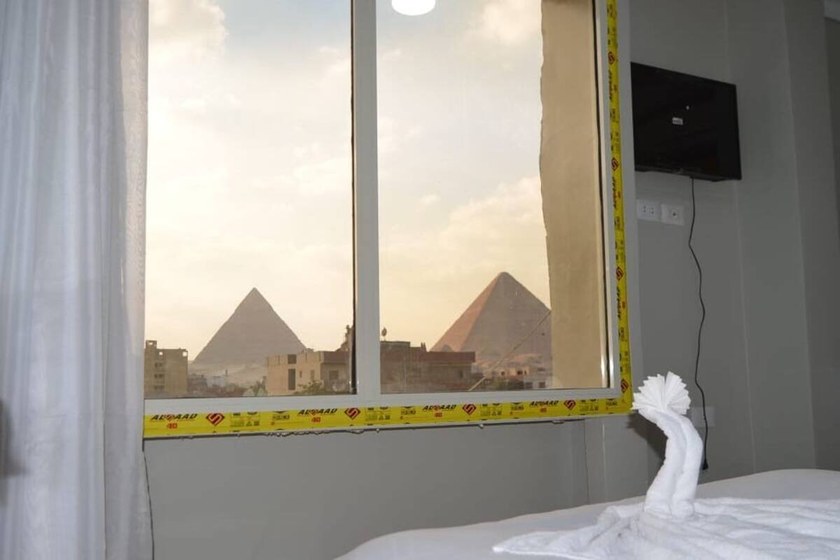 Una habitación de hotel con una cama, un ventanal con vistas a pirámides, un televisor montado en la pared y una toalla sobre la cama.