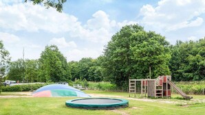 Un parque infantil con trampolín, tobogán y estructura de escalada en un área verde con árboles al fondo.