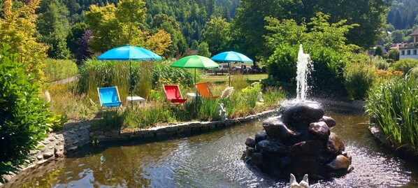 Un jardin avec un étang, une fontaine et un espace extérieur aménagé avec des tables et des parasols.