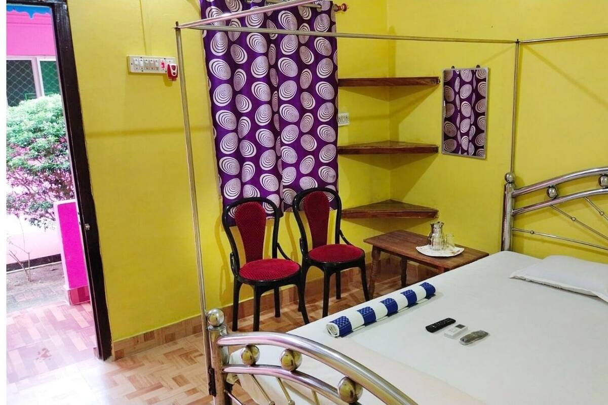 Een kamer met een bed, een kleine tafel, twee stoelen, een plank en een gordijn.