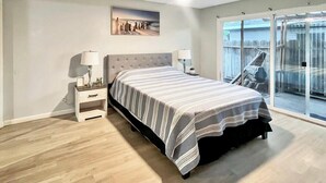 Un dormitorio con cama, mesita de noche, lámpara y un ventanal con vistas a un jardín.