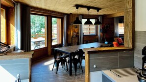 A modern kitchen with wooden ceiling beams, a dining area with a large table and chairs, and a view of the outdoors through large windows.