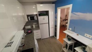 Une cuisine compacte avec des meubles blancs, un four à micro-ondes et un réfrigérateur. On y trouve un petit coin repas avec une table et des chaises, ainsi qu'un mur orné d'une fresque représentant des nuages.
