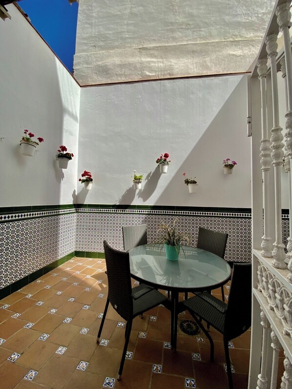 Un patio soleado con una mesa redonda de vidrio y sillas de mimbre negro, rodeado de baldosas estampadas y paredes blancas con macetas colgantes.