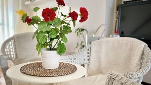 Un salon chaleureux avec un canapé en osier, une petite table ronde surmontée d’un set de table en fibres tressées et un vase blanc contenant des fleurs rouges.