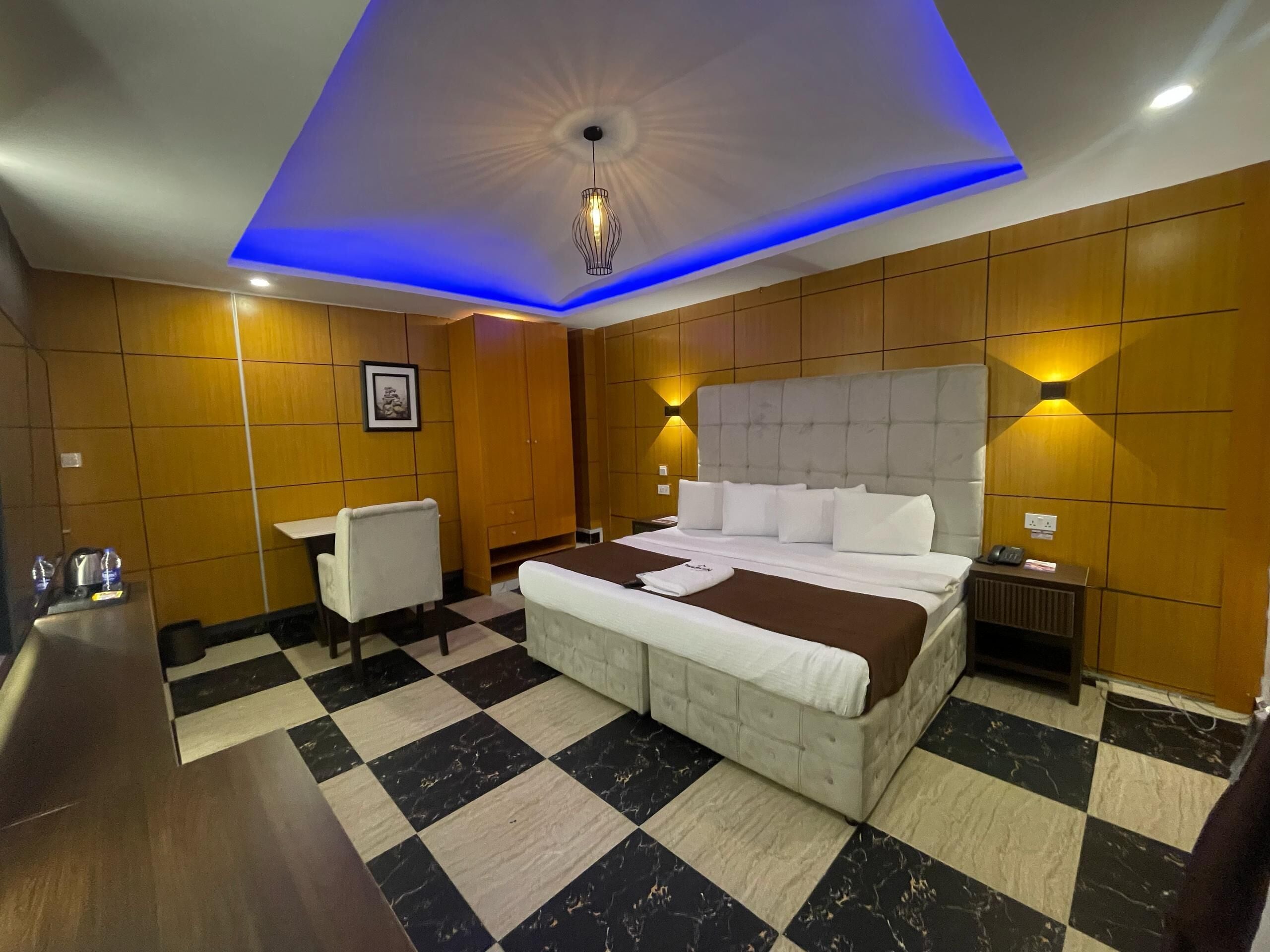 Quarto de hotel com chão xadrez, uma cama grande com roupa de cama branca, uma cômoda, uma escrivaninha com cadeira e um luminário fixado na parede.