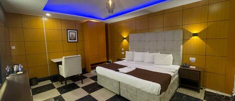 Quarto de hotel com chão xadrez, uma cama grande com roupa de cama branca, uma cômoda, uma escrivaninha com cadeira e um luminário fixado na parede.