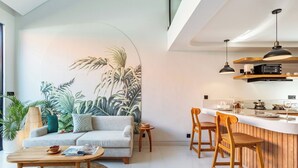 A modern living room with a large tropical mural, a sofa, a coffee table, and a dining area with wooden chairs and a kitchen counter. - Cove Anantaha Loft & Villa (Keroboken Kelod)