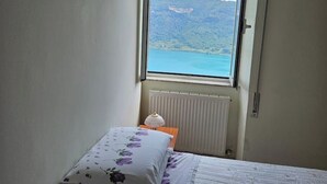 A bedroom with a bed, a nightstand, a lamp, and a window offering a view of a mountainous landscape and a body of water. - House with a view of Lake Nemi  (Genzano di Roma)