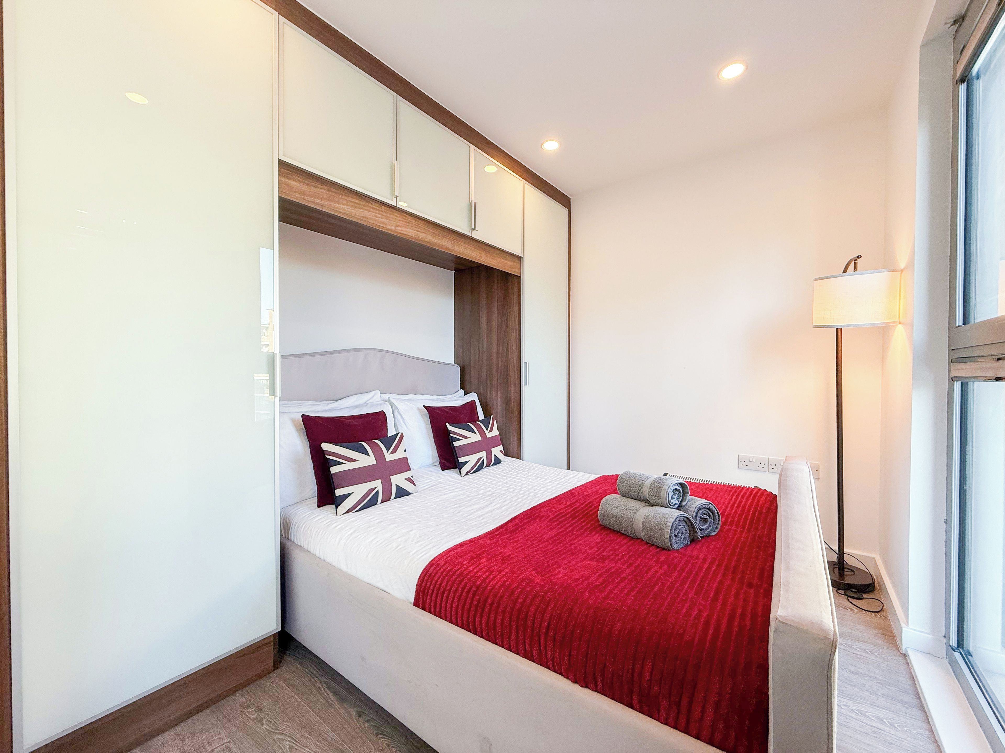 Een hotelkamer met een bed, een rode plaid, kussens met een Union Jack ontwerp, een lamp en een raam.