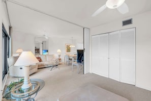 Uma sala de estar ampla com sofá, mesa de centro e televisão. Há um ventilador de teto, uma luminária e uma porta com venezianas.