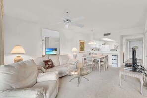 Un salon moderne comprenant un canapé, une table basse en verre, un coin repas avec une table et des chaises blanches, un coin cuisine avec des meubles blancs et un ventilateur de plafond.