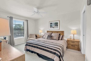 Un dormitorio con cama, dos mesitas de noche con lámparas, ventilador de techo, ventana con cortinas e imagen enmarcada en la pared.