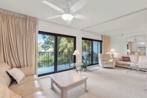 A spacious living room with a sofa, a coffee table, and a chair. There are large windows with curtains, a balcony, and ceiling fans.