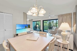 Ein modernes Hotelzimmer mit einem Holztisch, einem Fernseher, der eine tropische Strandlandschaft zeigt, einem weißen Sofa und einem Balkon mit Blick auf Palmen.