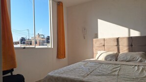 Ein Schlafzimmer mit einem Bett, einem Sessel, einem Fenster mit Vorhängen und Blick auf Gebäude.
