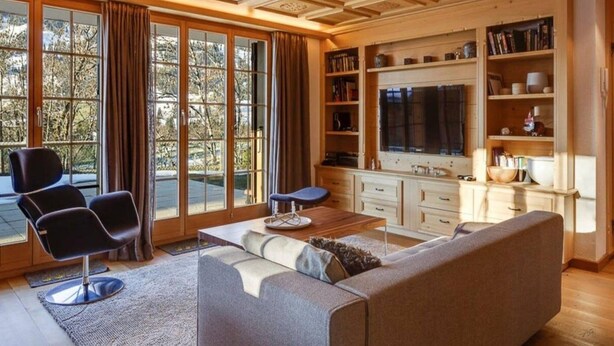 A spacious living room with a large window, a sofa, a coffee table, and a television. - Lush Apartment With Sauna and hot tub (Saanen)