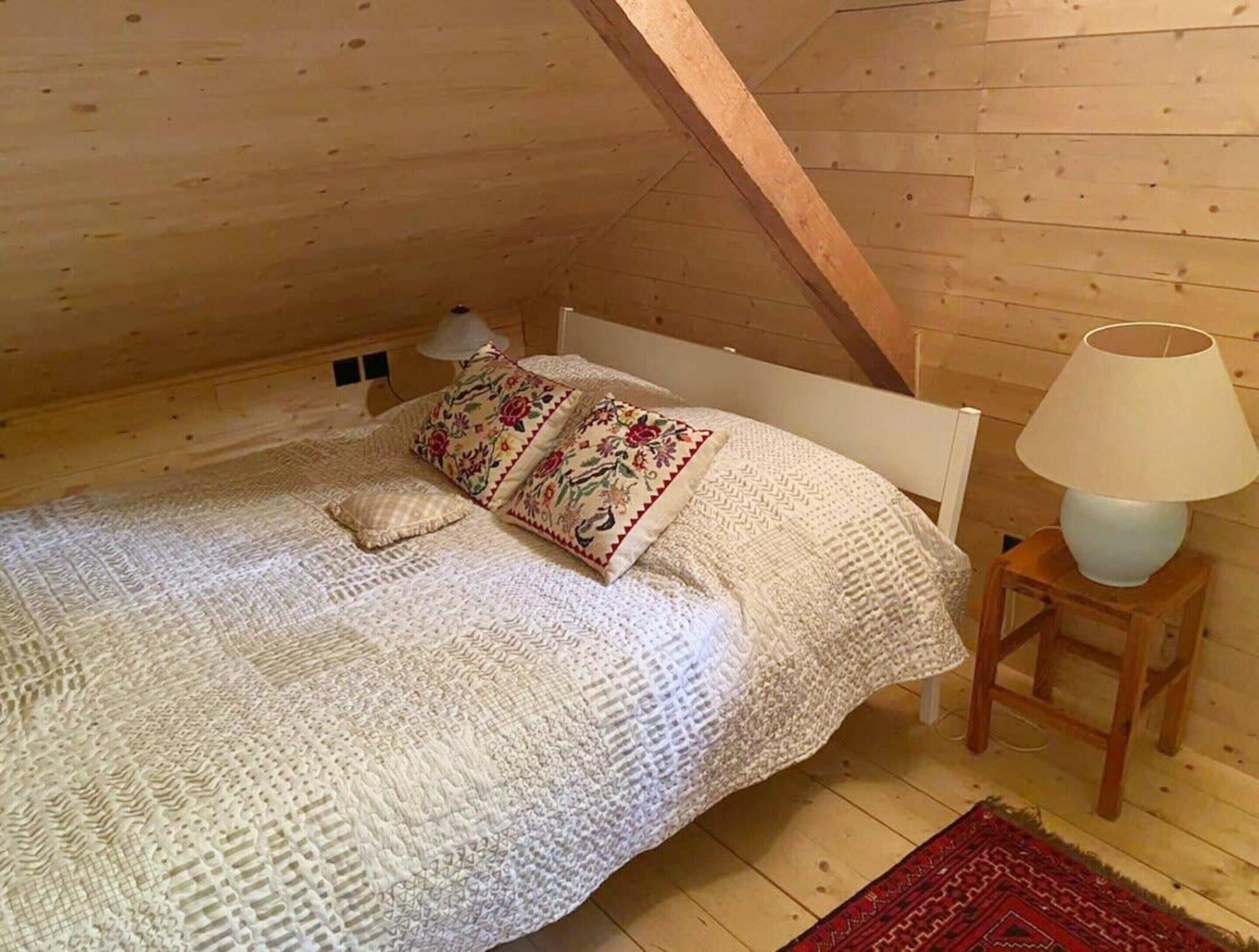 A cozy attic bedroom with a wooden sloped ceiling, a white bed with a patterned blanket, two decorative pillows, a bedside lamp, and a red rug on the wooden floor.