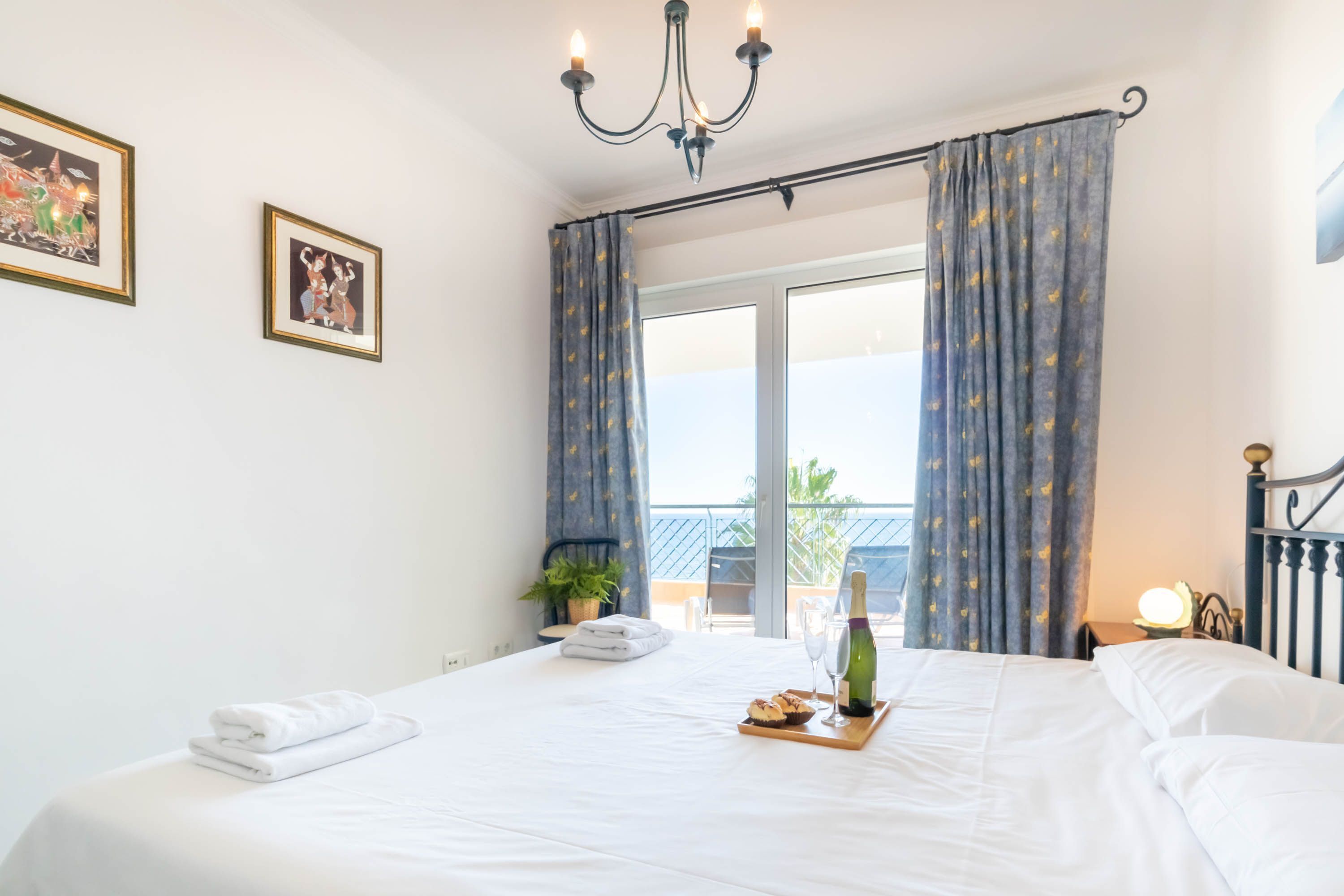 A hotel room with a large bed, a chandelier, a balcony with curtains, and a breakfast tray with a bottle and glasses.